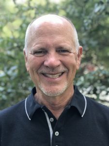 Smiling bald man in black polo shirt standing outdoors with green foliage background