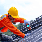 Worker in yellow hard hat and orange vest installing metal roof panels with power drill